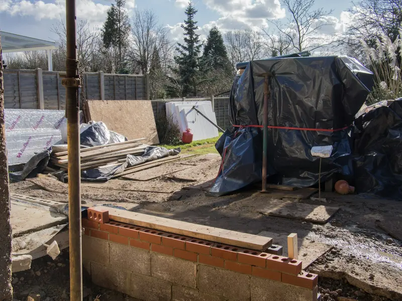 Residential foundation under construction with formwork, materials under tarps and a fenced yard